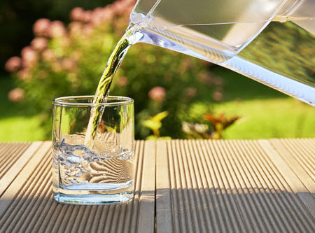 Pouring clear filtered water from a water filtration jug into a glass on the green summer garden background in a warm sunny summer day