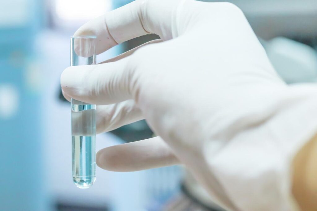 A gloved hand holding a glass testing tube filled with clear water.